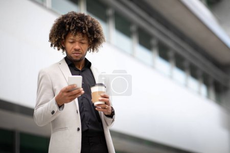 Foto de Empresario masculino afroamericano de mediana edad que sostiene café para llevar y usa el teléfono celular mientras camina al aire libre durante el descanso y pasa tiempo cerca del centro de oficinas, espacio para copiar - Imagen libre de derechos