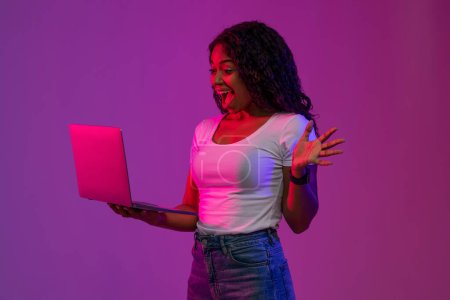 Téléchargez les photos : Cool Website. Portrait de femme noire excitée avec ordinateur portable dans les mains sous éclairage au néon, Lady afro-américaine réjouie célébrant le succès avec ordinateur portable, debout sur fond violet Studio - en image libre de droit