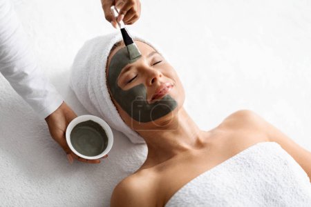 Photo for Anti-Aging Skincare. Cosmetologist applying clay mask on face of middle aged woman, beautiful mature female with towel on head lying on table, enjoying beauty tratments in spa salon, closeup - Royalty Free Image