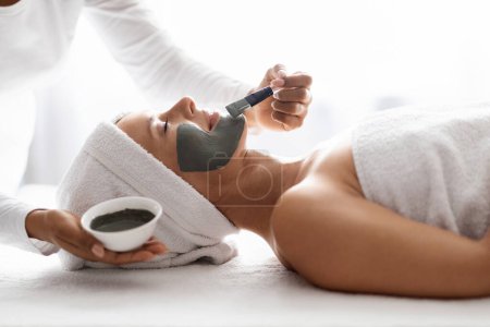 Photo for Side view of spa therapist applying clay mask on woman face, beautiful middle aged female enjoying skincare treatments at modern beauty center, lying on table and smiling, closeup shot - Royalty Free Image