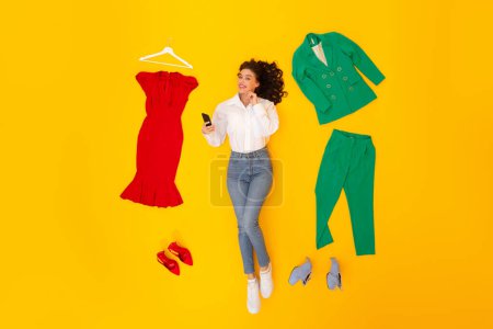 Photo for Happy Woman Shopping Online In Mobile Application Using Cellphone Lying Among Clothes And Shoes In Studio On Yellow Background, Smiling To Camera. Fashion And Technology Concept. Above View - Royalty Free Image