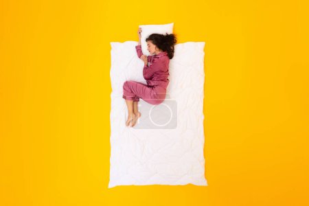 Téléchargez les photos : Une routine de sommeil saine. Dame paisible allongée sur une couverture et un oreiller portant un pyjama faisant la sieste en position foetale sur un arrière-plan de studio jaune, vue du dessus. Concept de loisirs et de confort - en image libre de droit