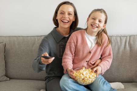Photo for Laughing happy european teen girl and millennial mom with remote control eating popcorn, watching tv in living room interior. Movie evening together, love, relationships, entertainment at home - Royalty Free Image