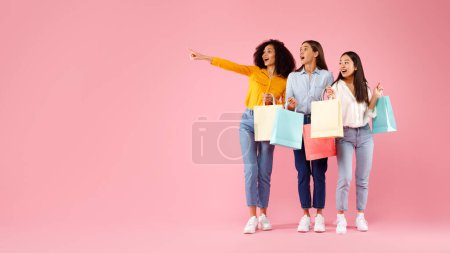 Photo for Three excited diverse ladies with colorful shopping bags pointing and looking aside, standing over pink studio background, panorama with copy space, full length - Royalty Free Image