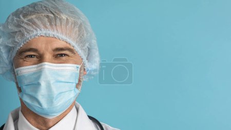 Téléchargez les photos : Portrait rapproché d'un homme d'âge moyen médecin chirurgien portant un masque facial et un filet à cheveux, posant sur fond de studio bleu, panorama avec espace de copie. Soins de santé, hôpital, concept de clinique - en image libre de droit