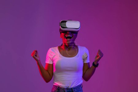 Téléchargez les photos : Jeune femme noire joyeuse portant des lunettes VR célébrant le succès dans la lumière au néon, joyeuse femme afro-américaine joyeuse acclamant gagner dans le jeu vidéo, debout sur fond violet, Copier l'espace - en image libre de droit
