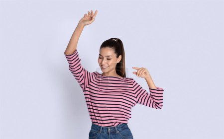 Photo for Cheerful cool carefree beautiful brunette young latin woman with ponytail wearing casual outfit dancing with closed eyes on grey studio background, raising hands up, having fun, copy space - Royalty Free Image