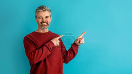 Photo for Cheerful smiling handsome mature caucasian man wearing red sweater pointing at free space for text or advertisement over blue studio background, showing nice offer, panorama - Royalty Free Image