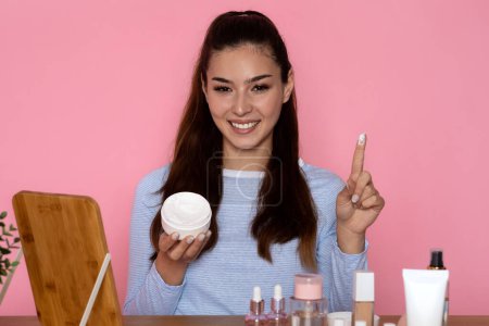 Photo for Excited happy pretty brunette young hispanic woman sitting at table full of diverse cosmetics, holding jar and showing her finger, checking brand new face cream, pink background, copy space - Royalty Free Image