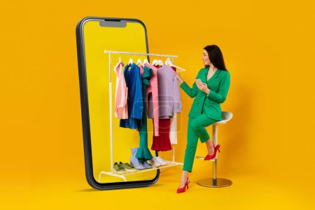 Photo for Young stylish woman sitting near huge cellphone and clothing rail using smartphone, buying clothes online via app over yellow studio background. Collage - Royalty Free Image