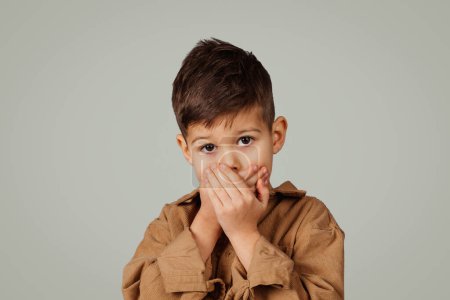 Foto de Impresionado niño de 6 años en casual cierra la boca con las manos aisladas sobre fondo gris. Miedo, estrés, problemas en la educación y la infancia, emociones y secretos - Imagen libre de derechos