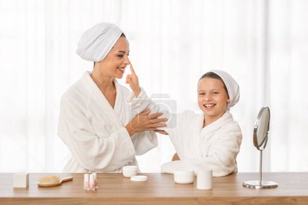 Photo for Mom And Daughter In Bathrobes Having Fun While Making Beauty Routine At Home, Happy Cute Preteen Girl And Her Mom Applying Moisturisung Cream And Laughing, Enjoing Time Together, Free Space - Royalty Free Image