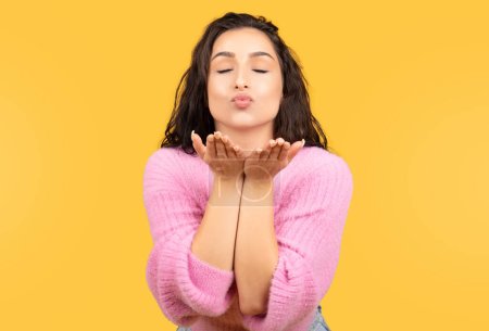 Téléchargez les photos : Femme arabe millénaire positive dans occasionnel avec les yeux fermés rend souffler baiser isolé sur fond jaune, studio. Amour, romance, soins de beauté, flirt, vente et style de vie, annonce et offre - en image libre de droit