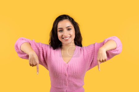 Photo for Positive millennial arab lady in casual wear showing fingers down to empty space isolated on yellow background, studio. Professional recommendation, advice, ad and offer, sale - Royalty Free Image