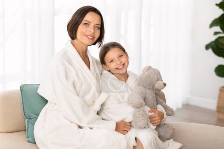 Photo for Mom And Child. Portrait Of Happy Young Mother And Little Daughter Wearing Bathrobes Relaxing On Couch At Home, Beautiful Family Resting After Spa Treatments, Female Kid Holding Teddy Bear - Royalty Free Image