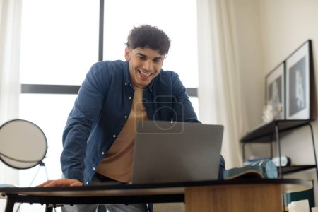Téléchargez les photos : Heureux homme du Moyen-Orient travaillant sur ordinateur portable debout au bureau moderne. Joyeux étudiant Guy travaillant et apprenant en ligne. Homme bénéficiant d'un travail indépendant et à distance. Internet et technologie - en image libre de droit