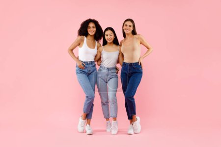 Téléchargez les photos : L'amitié des filles. Plan complet de trois femmes multiraciales heureuses étreignant et souriant à la caméra, posant debout sur fond de studio rose, espace libre - en image libre de droit