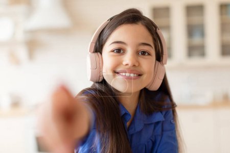 Téléchargez les photos : Portrait De Joyeux Brunette Preteen Girl portant des écouteurs écoutant de la musique en ligne, étirant la main à la caméra assis à la maison. Gadgets et Loisirs Concept. Focus sélectif - en image libre de droit