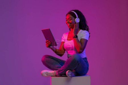 Téléchargez les photos : Gai noir femelle assis sur Gros cube dans néon lumière et en utilisant tablette numérique, heureux jeune afro-américaine écoute de la musique dans les écouteurs sans fil tout en se reposant avec moderne gadget - en image libre de droit