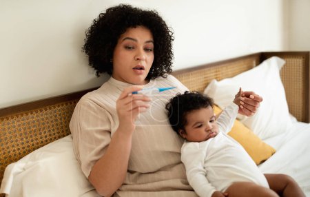 Photo for Worried sad shocked young african american mother with small child looks at thermometer, baby suffering from fever in bedroom interior. Parenting care, treatment, child health care at home - Royalty Free Image
