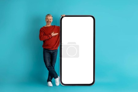 Photo for Cheerful smiling handsome middle aged man in casual outfit showing nice online offer on huge cell phone screen, mockup for advertisement over blue studio background, copy space - Royalty Free Image