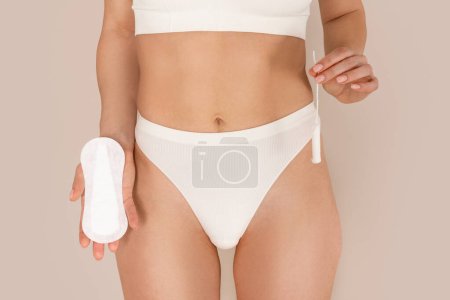 Photo for Female Period and Intimate Hygiene. Closeup cropped view of young woman in white underwear holding tampon vs clean menstrual pad near hips standing isolated on beige studio background - Royalty Free Image