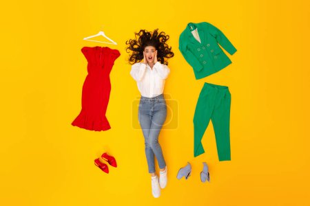 Photo for Omg, Shopping Offer. Shocked Shopaholic Lady Touching Face Lying Among Outfits Choosing Female Clothes And Shoes Posing Over Yellow Background, Studio Shot. Great Sales Concept - Royalty Free Image