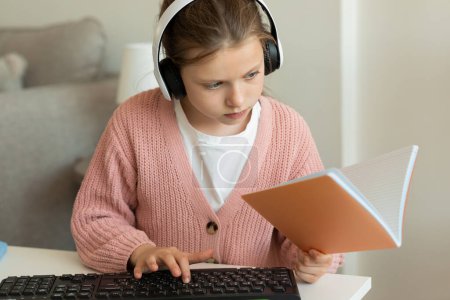 Photo for Busy european teenager girl in wireless headphones does homework and reads book, typing on computer in living room interior, close up. Study, knowledge, new normal remotely at home - Royalty Free Image