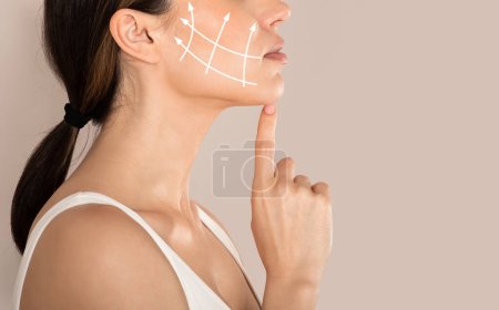Cropped of young woman with clean fresh skin touching her chin, antiaging concept. Side view of unrecognizable lady in white top with lifting arrows on cheek, beige studio background, copy space