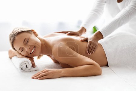 Téléchargez les photos : Belle dame d'âge moyen recevant un massage relaxant du dos par un thérapeute professionnel dans un salon de spa, gros plan d'une jolie femme mature bénéficiant d'un traitement de bien-être dans un studio de beauté, vue latérale - en image libre de droit