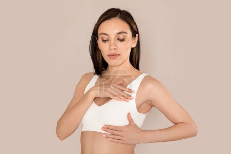 Photo for Breast Self-Exam, BSE. Beautiful young woman wearing underwear white top checking her breast on beige studio background. Concerned lady make screening, cancer prevention concept - Royalty Free Image