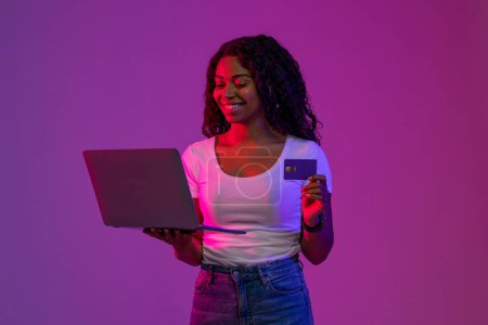 Photo for Happy Black Female Making Online Shopping With Laptop And Credit Card, African American Woman Standing In neon Light Over Purple Background, Enjoying Internet Purchases And E-Commerce, Free Space - Royalty Free Image