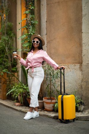 Téléchargez les photos : Positif jeune touriste femelle millénaire noir dans le chapeau et les lunettes de soleil, valise jaune avec tasse à café profiter de l'aventure seule en ville, verticale, pleine longueur. Vacances et voyages en plein air, week-end - en image libre de droit