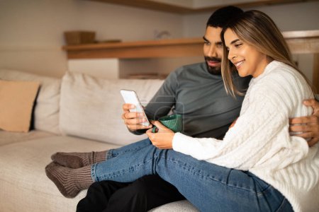 Photo for Happy millennial arabic husband hug european wife, looking at smartphone, have video call, enjoy romantic relationships in living room interior. App for communication remotely at home - Royalty Free Image