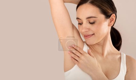 Photo for Closeup studio shot of young smiling beautiful woman wearing white top touching her fresh hairless armpit, isolated on beige studio background, panorama with copy space. Hyperhidrosis treatment - Royalty Free Image