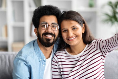 Photo for Happy cheerful cute beautiful young indian couple bearded guy wearing eyeglasses and long-haired lady taking selfie together at home, resting on couch, happy moments, closeup - Royalty Free Image