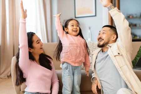 Téléchargez les photos : Famille asiatique joyeuse de trois avec une petite fille levant la main votant et jouant à la maison. Parents et enfants s'amusent ensemble le week-end. Happy Parenthood Concept - en image libre de droit