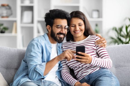 Photo for Relaxed cheerful beautiful young multicultural couple middle eastern guy and indian woman sitting on sofa at home, using smartphone and smiling, shopping online, surfing on Internet, copy space - Royalty Free Image