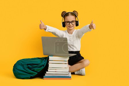 Photo for Happy Little Schoolgirl In Headphones Using Laptop Computer And Showing Thumbs Up At Camera, Cute Preteen Female Child Sitting With Book Stack Over Yellow Background, Advertising Educational Offer - Royalty Free Image