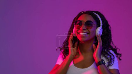 Téléchargez les photos : Femme afro-américaine joyeuse portant des lunettes de soleil élégantes écoutant de la musique dans les écouteurs sans fil, souriant heureux noir femelle debout dans la lumière au néon sur fond violet Studio, Copier l'espace - en image libre de droit