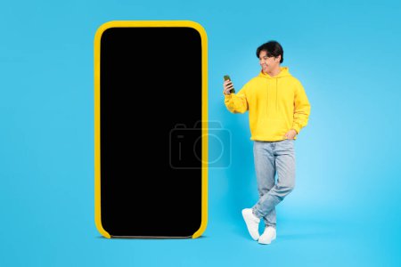 Photo for Mobile Offer. Japanese Teenager Guy Using Phone Posing Near Big Smartphone With Blank Screen Standing In Studio Over Blue Background. Cheerful Teen Boy Advertising New App. Mockup, Collage - Royalty Free Image