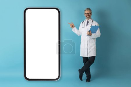 Photo for Positive happy smiling handsome grey-haired mature doctor with medical chart in his hand pointing at big phone with white empty screen, blue studio background, mockup, telemedicine concept - Royalty Free Image