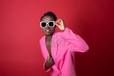 Photo for Fashion Shot Of Young Black Woman Wearing Pink Jacket And Stylish Sunglasses Standing Over Red Background, Gramorous African American Female Model Looking At Camera While Posing In Studio, Copy Space - Royalty Free Image
