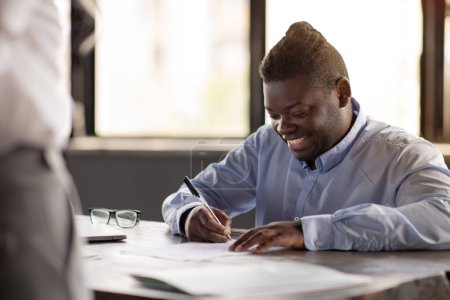 Téléchargez les photos : Homme d'affaires afro-américain joyeux signant des documents de contrat de travail pendant l'entretien d'emploi avec le directeur des RH assis dans le bureau moderne. Documentation et paperasse. Focus sélectif - en image libre de droit