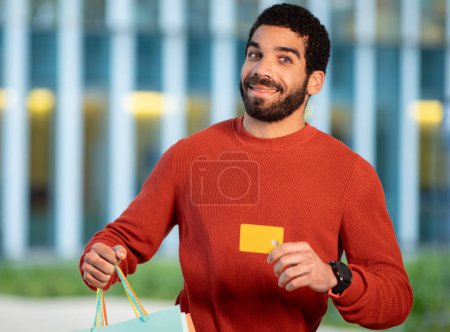 Photo for Shopping Offer. Cheerful Bearded Brunette Man Showing His Credit Card And Paper Shopper Bags, Advertising Bank Payment Service Smiling To Camera Standing Outdoors. Selective Focus - Royalty Free Image