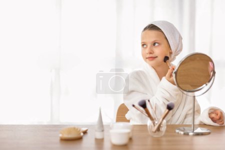 Foto de Linda niña aplicando maquillaje mientras está sentada en el tocador en casa, feliz niña preadolescente usando albornoz usando cepillo Blusher y mirando hacia un lado en el espacio de copia, disfrutando de la rutina de belleza - Imagen libre de derechos