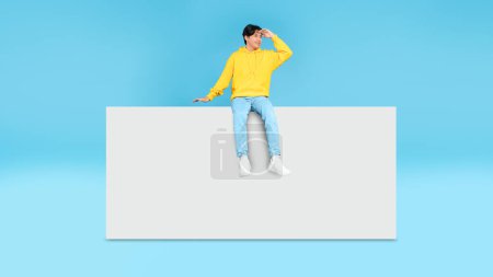 Photo for Cheerful Korean Teenager Guy Sitting On Blank Paper Poster Looking Aside Holding Hand At Forehead Advertising Great Offer Over Blue Studio Background. Look There Concept. Panorama With Copy Space - Royalty Free Image