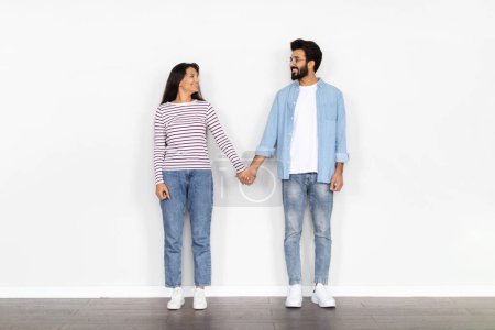 Cheerful happy loving beautiful young eastern spouses bearded man and long-haired woman wearing casual posing at new empty house, holding hands and smiling to each other, white wall background