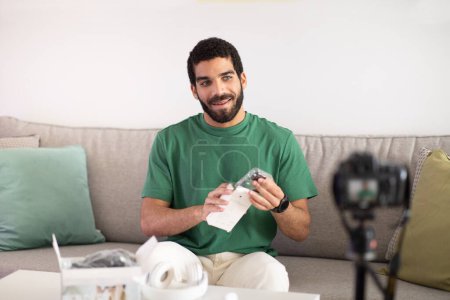 Photo for Cheerful young middle eastern man blogger shows headphones, shooting video on camera, enjoy work in room interior. Overview blog, online shopping review, work remotely at home - Royalty Free Image