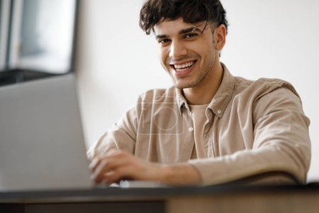 Téléchargez les photos : Homme arabe heureux utilisant ordinateur portable dactylographie et travail assis en ligne à la table dans le bureau moderne. Jeune homme d'affaires souriant à la caméra posant au travail à l'intérieur. Focus sélectif - en image libre de droit